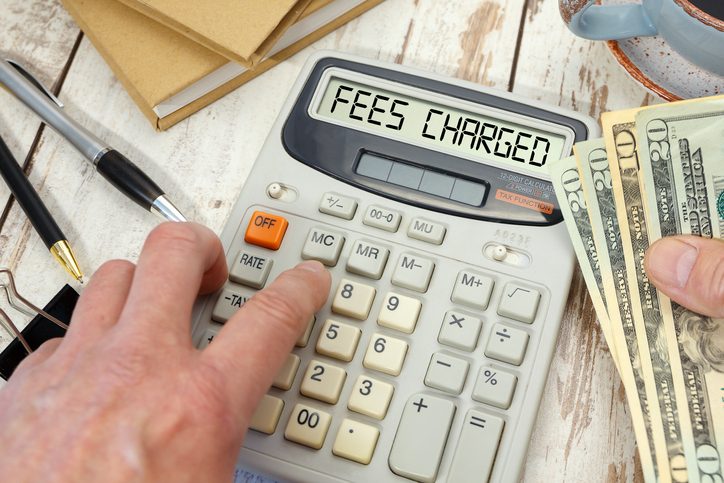 Money in hand next to a calculator and office supplies. FEES CHARGED words on calculator.